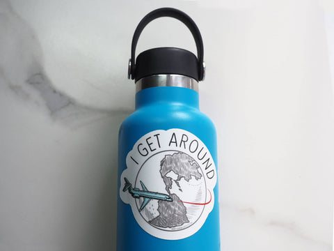 A round white sticker with a sketched world map and an airplane, and the text 'I GET AROUND' on it.