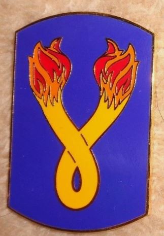 Shield-shaped emblem with a yellow ribbon and red flames on a blue background