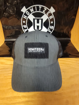 A black Howitzer branded trucker hat with a velcro patch on the front, displayed on a flat surface.