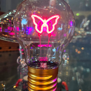 A neon light bulb with a butterfly , moon or star design illuminated in pink color, attached to a gold-colored base.