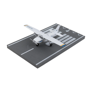 A miniature model of a white Cessna airplane with blue and yellow accents, displayed on a gray miniature runway base with white markings.