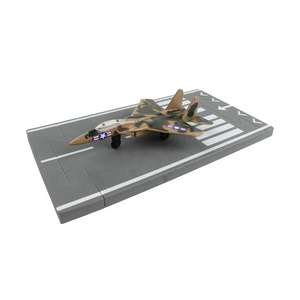 A toy model of a military fighter jet in a desert camouflage pattern, displayed on a gray miniature aircraft carrier deck against a plain white background.