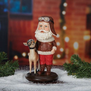 A figurine of Santa Claus wearing aviator gear and a bobblehead of a reindeer accompanying him, placed on a base of snowy ground with Christmas decorations in the background.