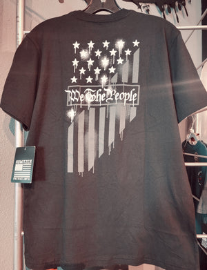 A black youth-sized t-shirt displayed on a hanger, featuring a flag design with white stars and the text 'We The People' on the front.