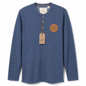 A blue long-sleeve henley shirt with a small brown circular patch on the chest and a hanging paper brand tag, shown on a plain white background.