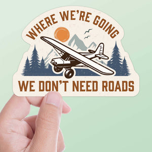 A die-cut vinyl decal featuring an airplane with the phrase 'Where we're going we don't need roads' against a mountain and pine forest backdrop.