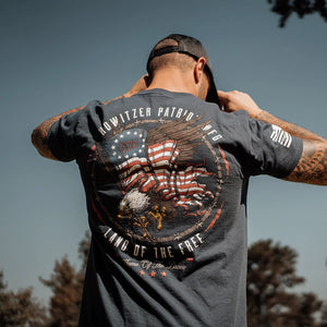 A person wearing a navy-colored short-sleeve t-shirt with a graphic that includes an eagle, the American flag, and the text 'Howitzer Patriot'. The shirt is turned back to show the front with the flag and eagle design.