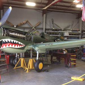 A P-40 Warhawk aircraft with a shark design on the mouth, parked inside a hangar with various tools and equipment around.