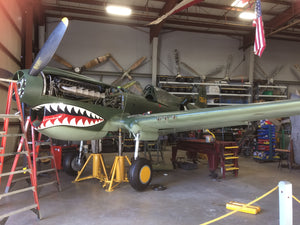 A P-40 Warhawk aircraft with a shark design on the mouth, parked inside a hangar with various tools and equipment around.
