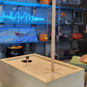 A wooden model of a Cessna airplane with a joystick, displayed on a tabletop at a museum-like setting with blurred objects in the background.