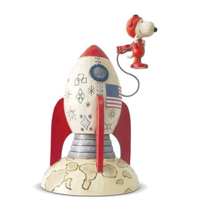 A figurine of Snoopy the astronaut, where Snoopy is depicted wearing a space suit and sitting in a rocket that is decorated with stars and an American flag.