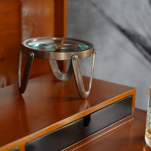 A brass desk magnifier with a glass base resting on a wooden surface.