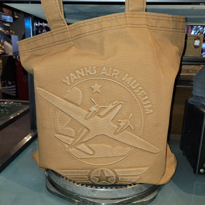 A beige canvas tote bag with embossed text and an airplane design, displayed on a glass shelf.