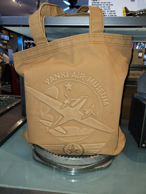 A beige canvas tote bag with embossed text and an airplane design, displayed on a glass shelf.